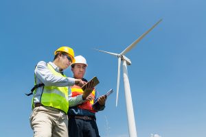 Engineer and Architect discuss over Digital Wireless Tablet and Clipboard while working at Wind Turbine Power Generator Tower site.