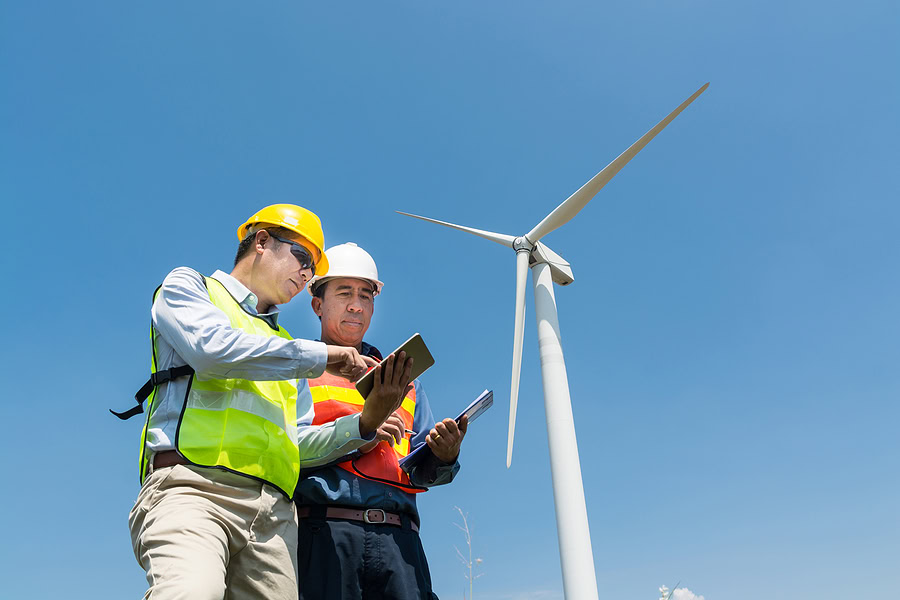 Engineer and Architect discuss over Digital Wireless Tablet and Clipboard while working at Wind Turbine Power Generator Tower site.