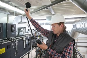 Technician testing air quality and flow using a device and wearing a white construction hat and dark vest.