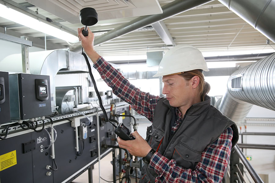 Technician testing air quality and flow using a device and wearing a white construction hat and dark vest.