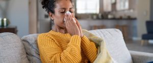 A person sitting on a couch wrapped in a blanket, holding a tissue to their nose.